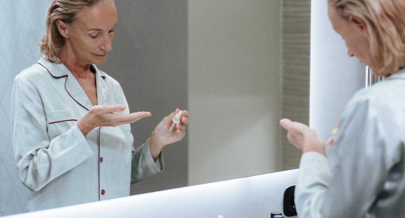 Woman in her fifties applying facial oil drops to her cheekbone in a clean bathroom mirror